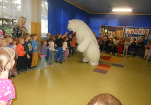 Dzieci stoja w kole i wyciągaja ręce do misia.