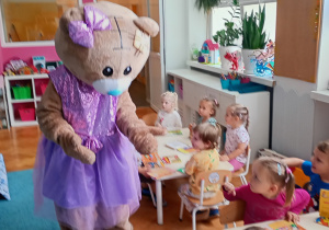 Miś przybija piątki z dziećmi siedzącymi przy stolikach, na ich twarzach widać radość i uśmiech