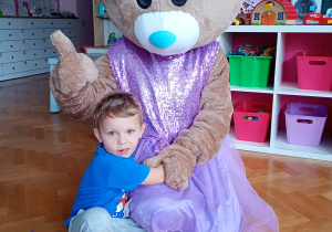 Mateusz pozuje do zdjęcia z misiem ubranym w fioletową spódniczkę i kokardkę