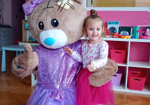 Emilka pozuje do zdjęcia z misiem ubranym w fioletową spódniczkę i kokardkę
