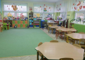 Sala grupy Żabki. Znajdują się w niej regały z zabawkami dla dzieci i niskie stoliki. Dywan i ściany są w kolorze zielonym. Na firankach wiszą dekoracje.