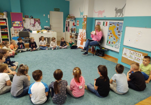 Dzieci siedzą w kole na dywanie oglądając obrazek pokazywany przez nauczycielkę (liczba "16")