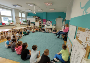 Dzieci siedzą w kole na dywanie, powtarzają słownictwo prezentowane przez nauczycielkę na podstawie kolorowych ilustracji