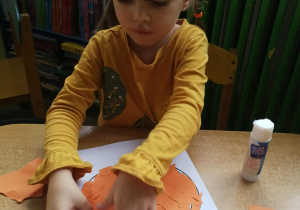 Lena nakleja kawałki pomarańczowej kartki na sylwetę dyni