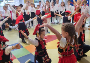 Dzieci ubrane w tradycyjne stroje tańczą krakowiaka (na planie dwóch kół chłopcy klęczą w wewnętrznym, dziewczynki stoją w zewnętrznym)