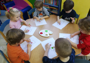 Dzieci przy niebieskim stole wystukują rytm piosenki "Kolorowe światła", tworząc kropki z farby na kartach z sygnalizatorami