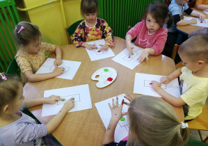 Dzieci przy zielonym stole wystukują rytm piosenki "Kolorowe światła", tworząc kropki z farby na kartach z sygnalizatorami