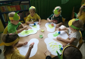 Dzieci przy żółtym stole stemplują pędzlami z gąbek umoczonymi w farbach sylwety gruszek