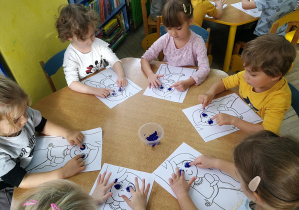 Dzieci przy niebieskim stoliku wystukują rytm piosenki "Oczy Jesieni" na ilustracjach palcami umoczonymi w farbach - dwie kropki w ustawieniu poziomym