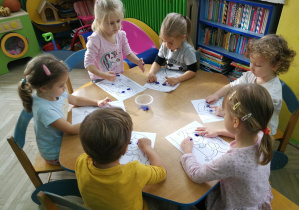 Przedszkolaki przy niebieskim stoliku wystukują rytm piosenki "Oczy Jesieni" na ilustracjach palcami umoczonymi w farbach
