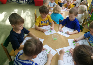 Przedszkolaki przy zielonym stoliku wystukują rytm piosenki "Oczy Jesieni" na ilustracjach palcami umoczonymi w farbach