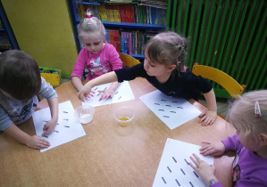 Dzieci przy żółtym stoliku naklejają makaron na kreskach narysowanych na karcie