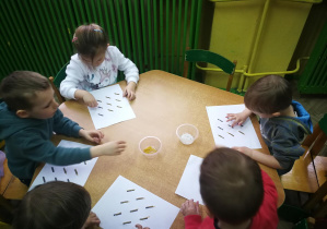 Dzieci przy zielonym stoliku naklejają makaron na kreskach narysowanych na karcie