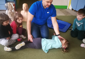 Ratownik medyczny pokazuje dzieciom w jaki sposób ułożyć poszkodowanego w pozycji bezpiecznej