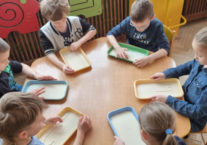 Dzieci przy żółtym stoliku piszą małą literę "l "palcem na tacce z manną