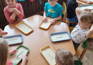 Dzieci przy zielonym stoliku piszą małą literę "l"palcem na tacce z manną