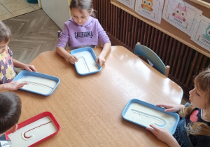 Dzieci przy niebieskim stoliku piszą małą literę "l" palcem na tacce z manną