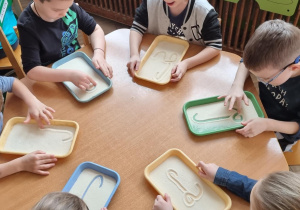 Dzieci przy żółtym stoliku piszą wielką literę "L "palcem na tacce z manną