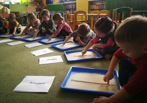 Dzieci rysują palcami na kaszy mannie linie pionowe w rytmie piosenki