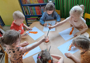 Dzieci przy żółtym stoliku malują pomarańczową farbą sylwety marchewek
