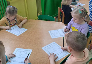 Dzieci przy zielonym stoliku rysują ołówkiem na rysunkach poziome linie od lewej do prawej w rytmie piosenki "Zaczarowany ołówek"