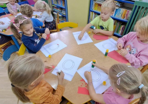 Dzieci przy żółtym stoliku wyklejają czerwoną wydzieranką wzory litery "o"