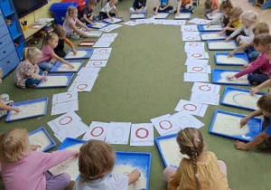 Dzieci kreślą litery "o" na tackach z kaszą manną do piosenki "Ola"