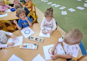 Dzieci przy niebieskim stoliku kreślą literę "o" na kartce, śpiewając piosenkę "Ola"