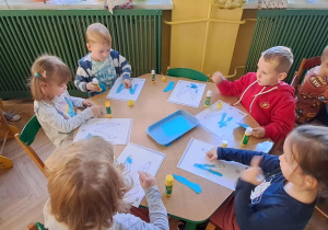 Dzieci przy zielonym stole wyklejają niebieską wydzieranką wzory litery "M"