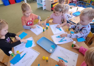 Dzieci przy żółtym stole wyklejają niebieską wydzieranką wzory litery "M"