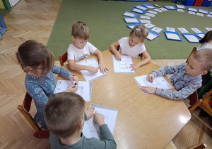 Dzieci przy czerwonym stole kreślą litery "E" na kartkach, jednocześnie śpiewając piosenkę "Ewelina"