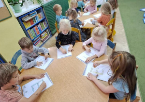 Dzieci przy niebieskim stole kreślą litery "E" na kartkach, jednocześnie śpiewając piosenkę "Ewelina"