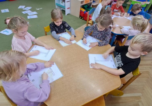 Dzieci przy żółtym stoliku kreślą literę "T" w rytmie piosenki "Tomasz" na kartkach kredkami świecowymi