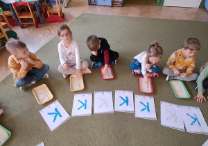 Pięciolatki kreślą litery "K" na tackach z kaszą manną, śpiewając piosenkę "Krystyna"