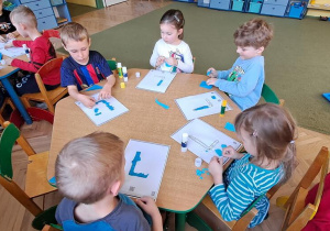 Dzieci przy zielonym stole wyklejają litery "L" niebieską wydzieranką