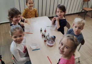 Dzieci siedzą przy stolikach z przygotowanymi elementami do tworzenia slime