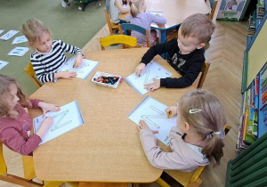 Dzieci przy żółtym stole kreślą litery "N" na kartkach kredkami świecowymi w rytmie piosenki "Natalia"
