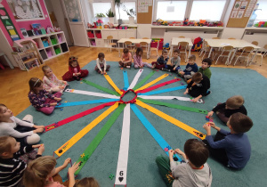 Dzieci siedzą na dywanie, na środku koła rozłożony jest kolorowy wiatrak matematyczny