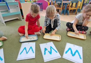 Oliwka, Marysie i Remik rysują litery "W" na tackach z kaszą w rytmie piosenki "Weronika"