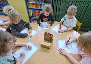 Dzieci przy żółtym stole piszą litery "Ł" kredkami na kartkach do piosenki "Łukasz"