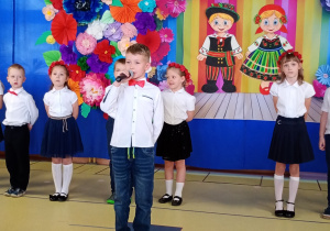 Wojtek recytuje wiersz, w tle stoją dzieic ubrane na biało- granatowo