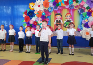 Aleks recytuje wiersz, w tle stoją dzieic ubrane na biało- granatowo
