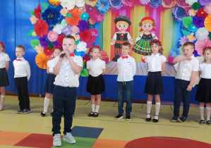 Oskar recytuje wiersz, w tle stoją dzieic ubrane na biało- granatowo