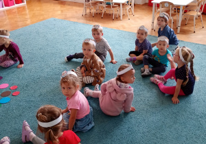 Dzieici siedzą na dywanie, mają białe opaski na głowie.