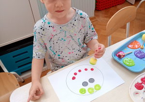 Chłopiec siedzi przy stoliku, ma na głowie białą opaskę. Wykonuje pracę plastyczną z kolorowych kropek.