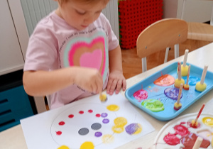 Dziewczyna w kolorowej bluzce siedzi przy stoliku. Wykonuje pracę plastyczną farbami z kolorowych kropek