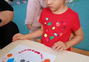 Dziewczyna w czerwonej bluzce siedzi przy stoliku. Wykonuje pracę plastyczną farbami z kolorowych kropek