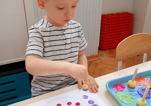 Chłopiec w bluzce w paski ssiedzi przy stoliku. Wykonuje pracę plastyczną farbami z kolorowych kropek