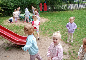 Dzieci w kolowych ubraniach bawią się w rozsypce w ogrodzie przedszkolnym. W tle czerwona zjeżdżalnia