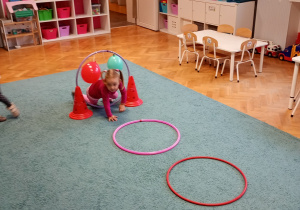 Dziewczynka w różowej bluzce pokonuje tor przeszkód na niebieskim dywanie – z zaangażowaniem i radością uczestniczy w aktywności ruchowej